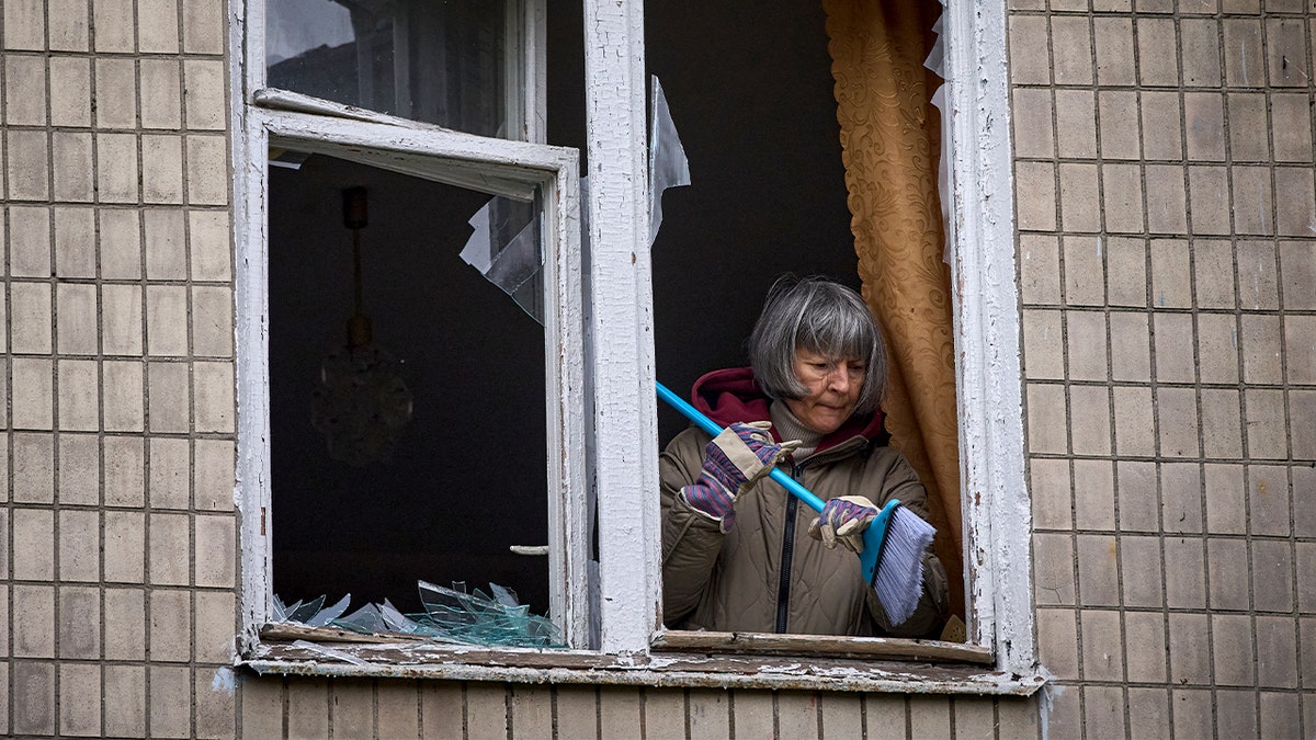 A resident clears broken glass from a window inside a damaged apartment following an aerial strike.