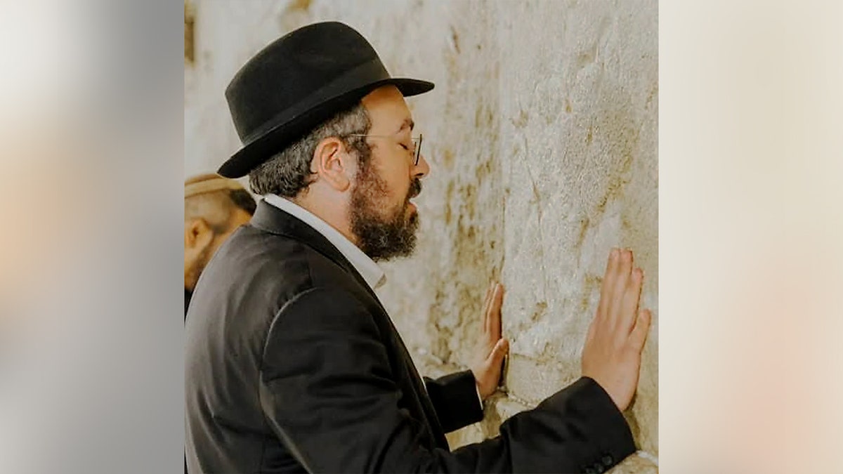 Man in a black hat and suit prays with his hands on a stone wall.