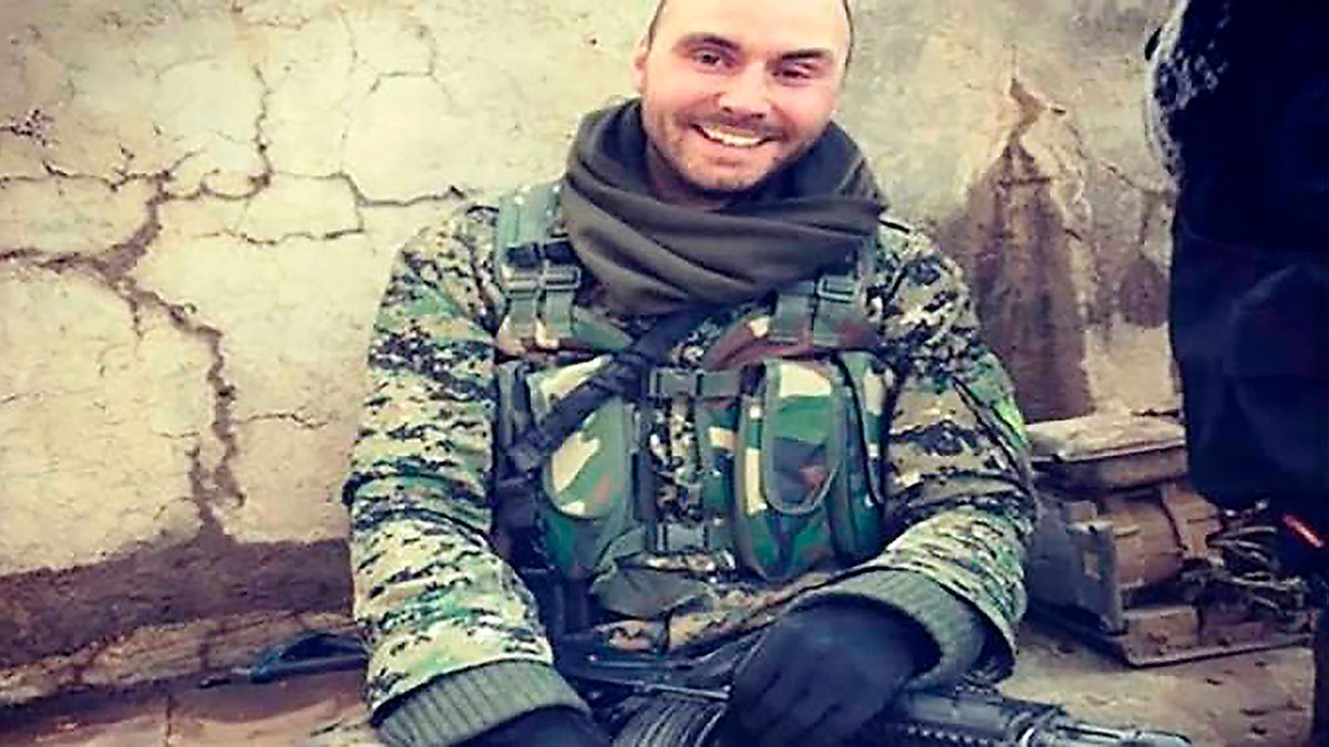 Man in camouflage uniform sits against a cracked wall while holding a rifle.