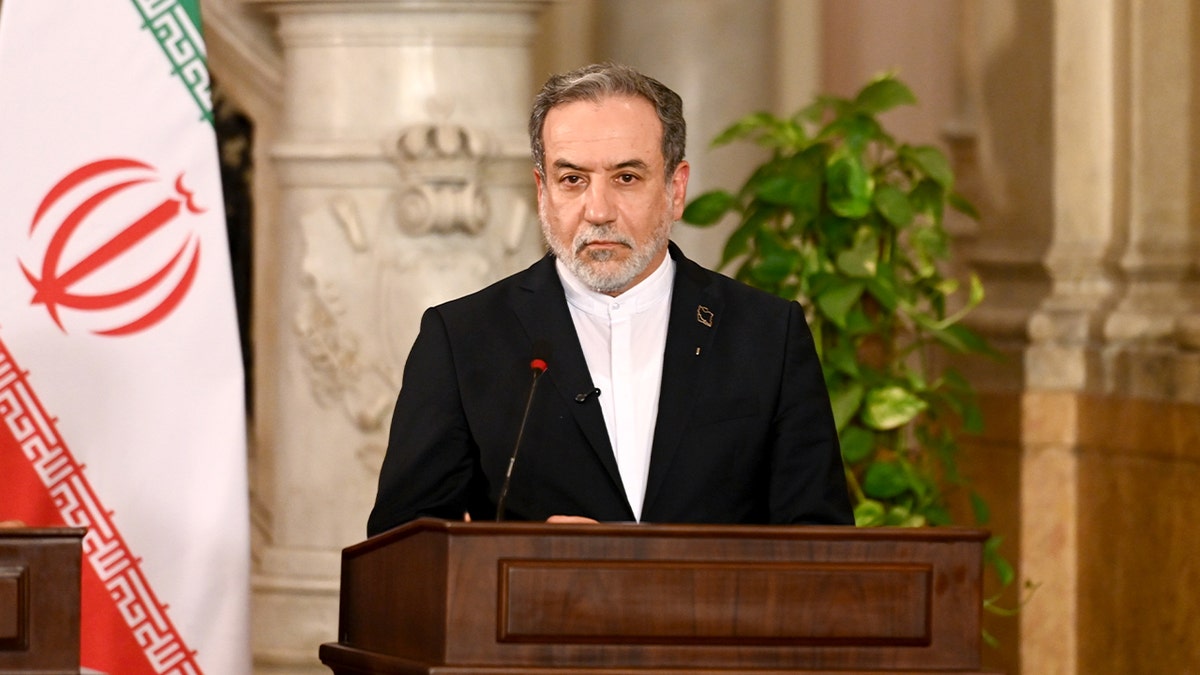 An Iranian official stands at a podium during a formal press conference, with national flags positioned behind him.