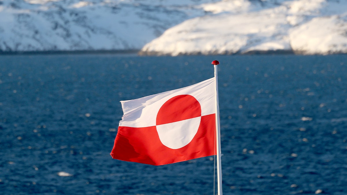 A red-and-white flag waves on a pole against a cold Arctic sky in a coastal city.