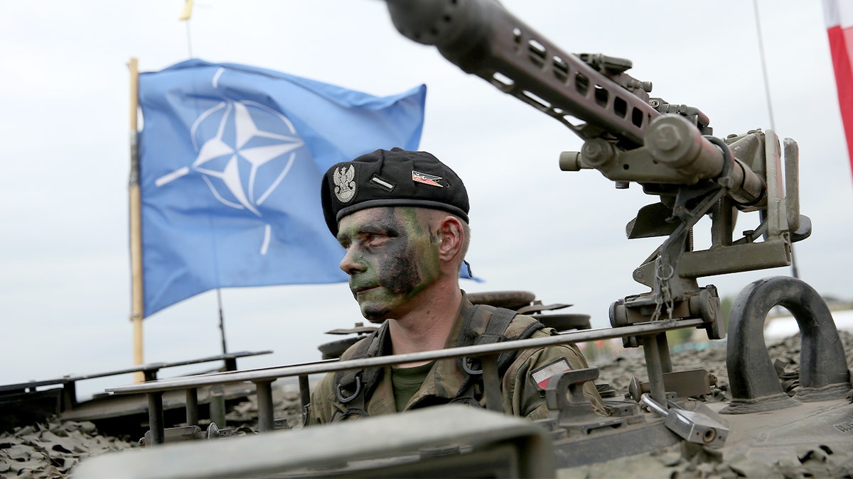A Polish soldier sits inside a military tank with a NATO flag visible in the background.