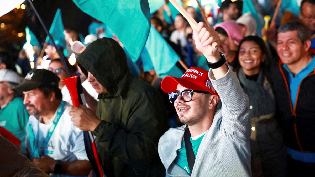 A man in a red political cap looks on with an animated expression inside a crowded indoor venue.