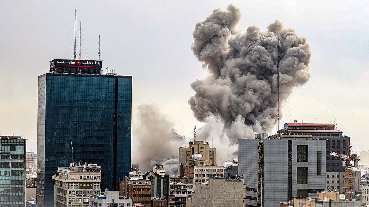Tehran city skyline with dark smoke rising from a distance under a hazy sky.