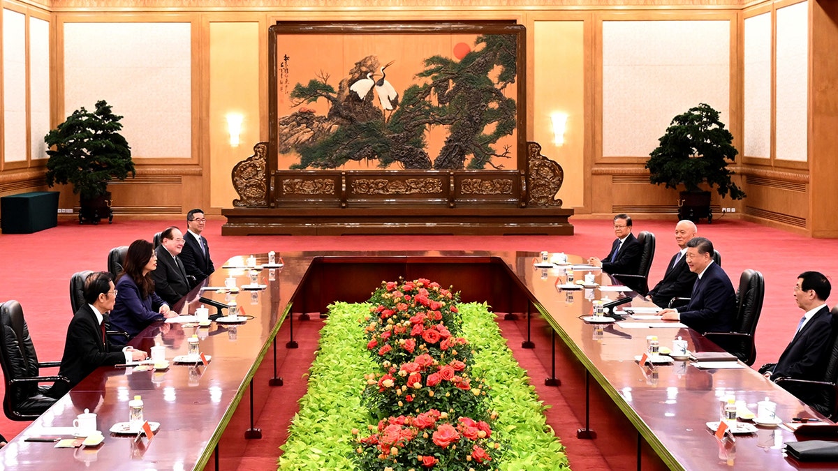 Dignitaries line two tables facing each other with a tapestry in the background and florals in between the two tables.