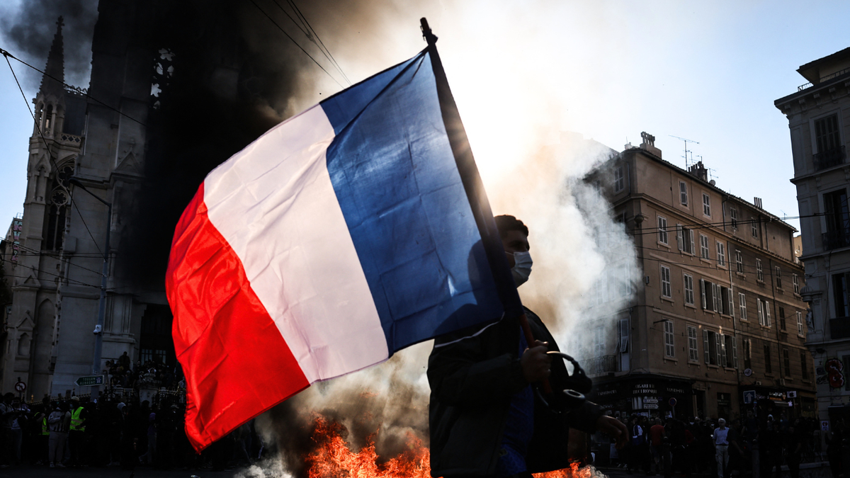 A protester waving a French national flag walks past a fire during clashes prior to the start of a demonstration to protest against the "high cost of living", in Marseille, southern France, on October 17, 2026.