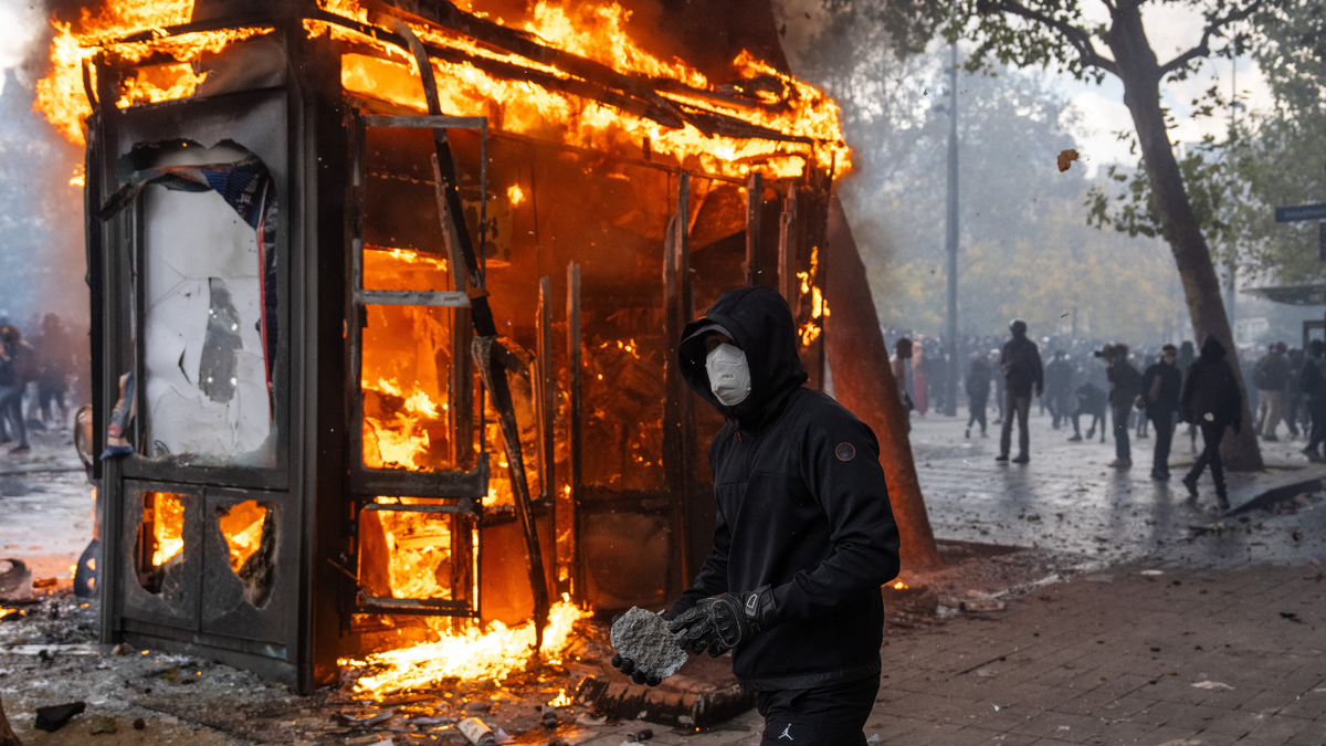 A protester holds a rock to throw at police as a kiosk burns during a demonstration near Place de la République, as the the Gilets Jaunes join students for a weekend of action on October 17, 2026 in Paris, France.